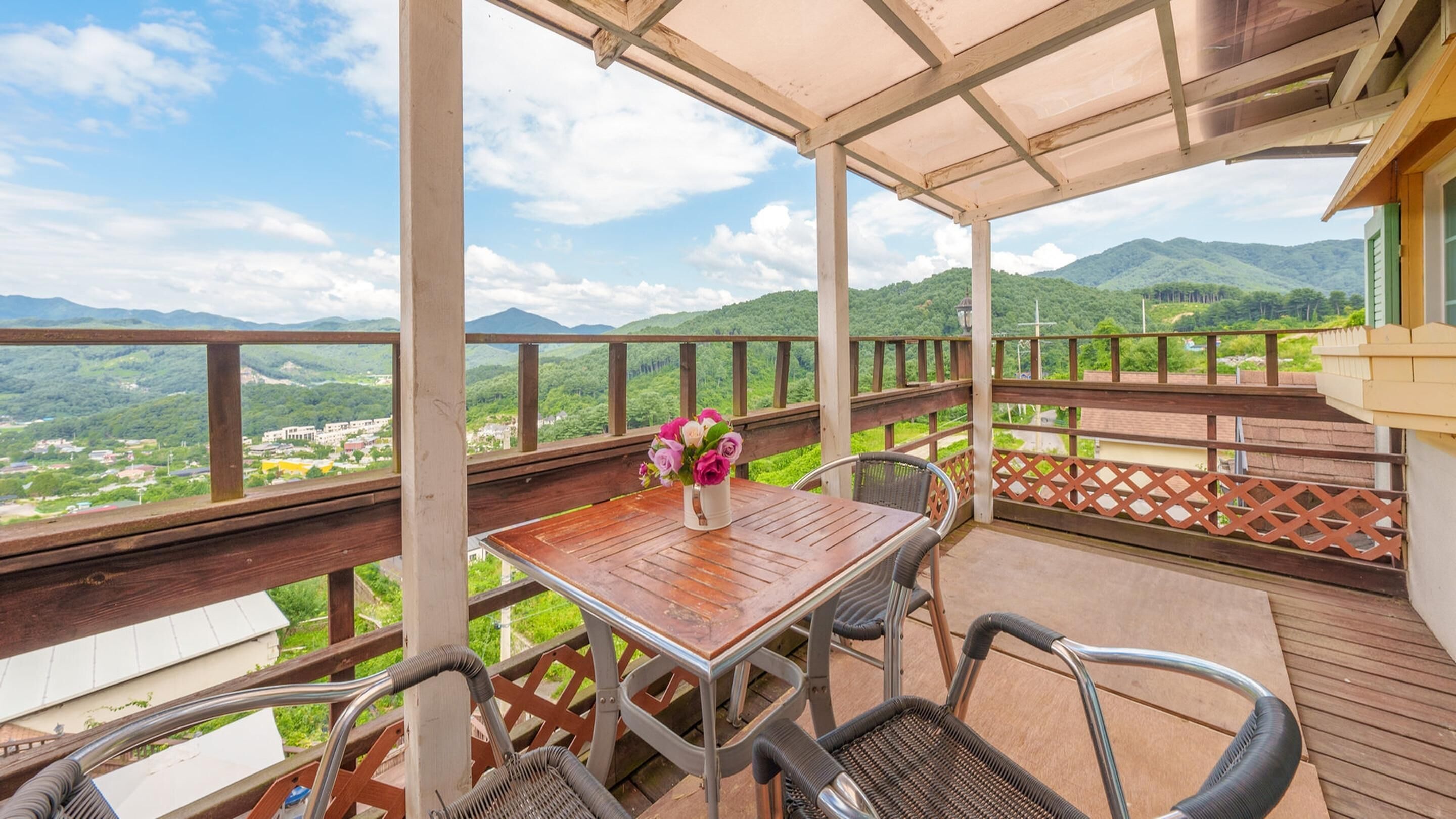 A covered outdoor area with a wooden table and chairs, a vase of flowers, and a view of green hills and mountains.