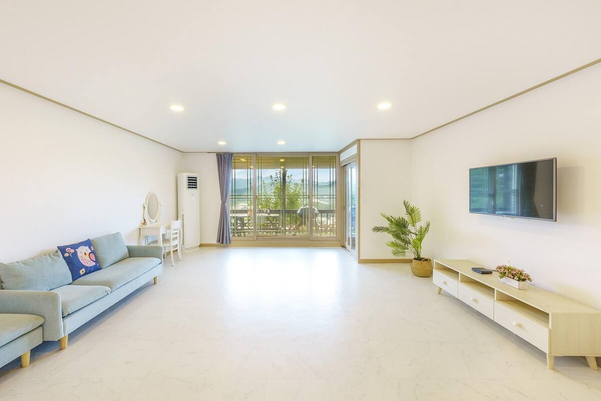 A spacious living room with a sofa, a TV, a plant, and a balcony with a view.
