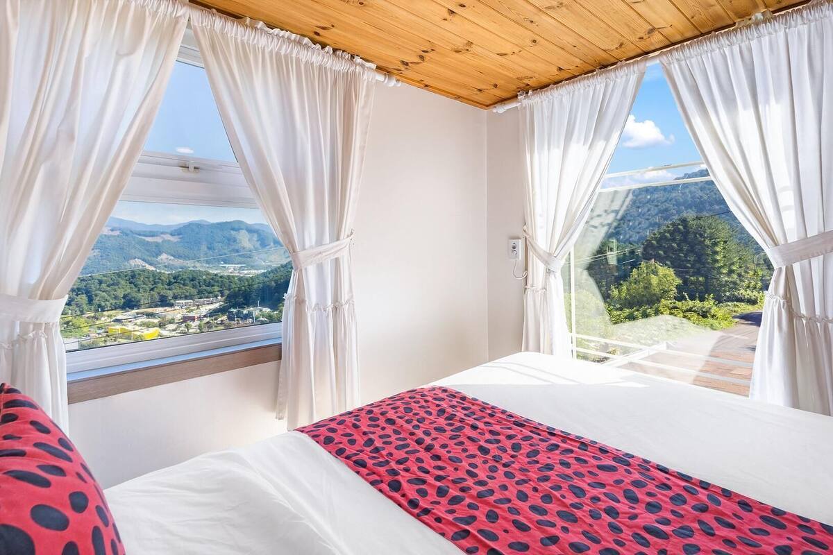 A bedroom with a bed, a view of a mountainous landscape, and white curtains.