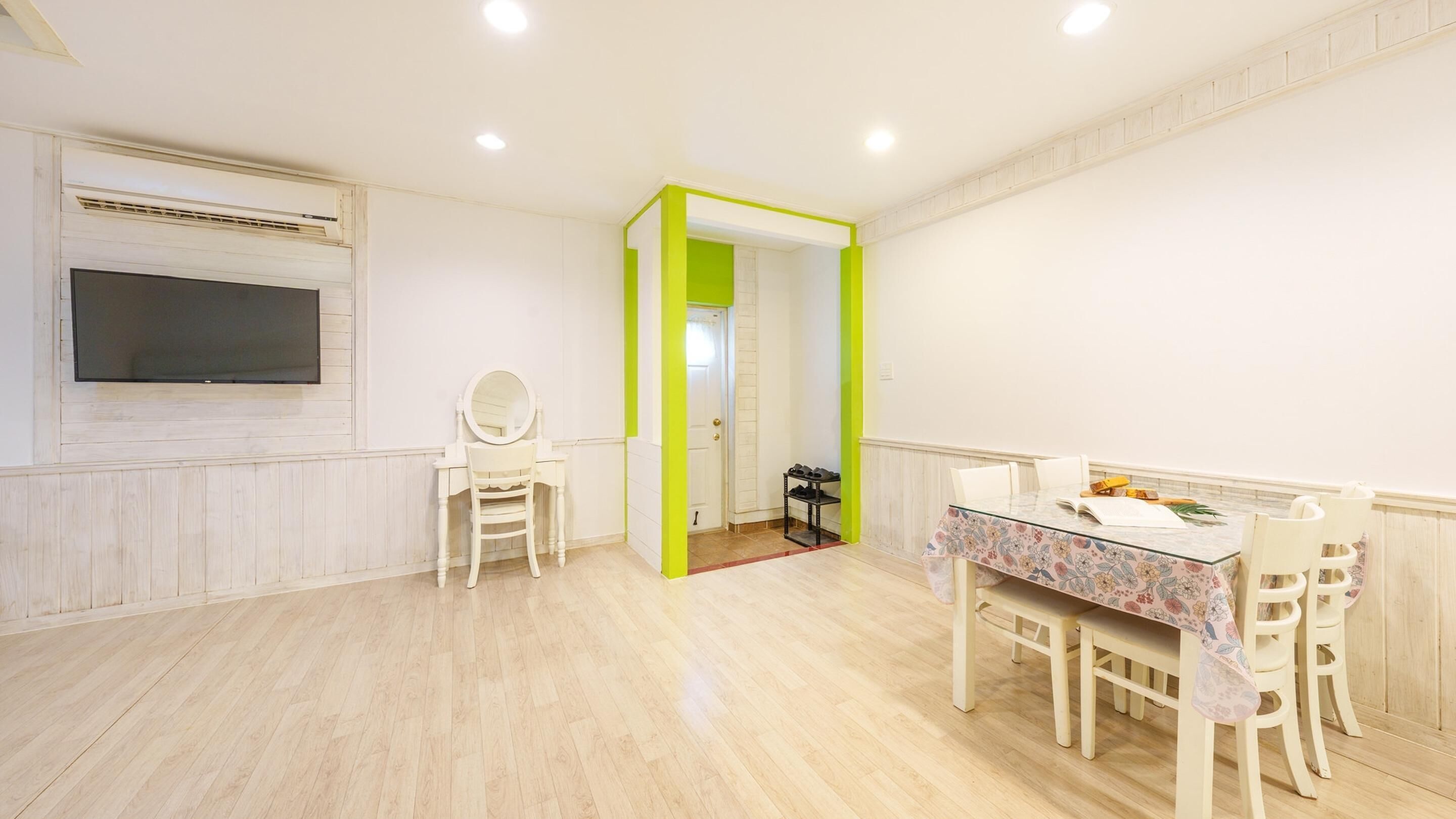 A bright room with a wooden floor, a white vanity with a mirror, a dining table with chairs, and a door leading to another room.