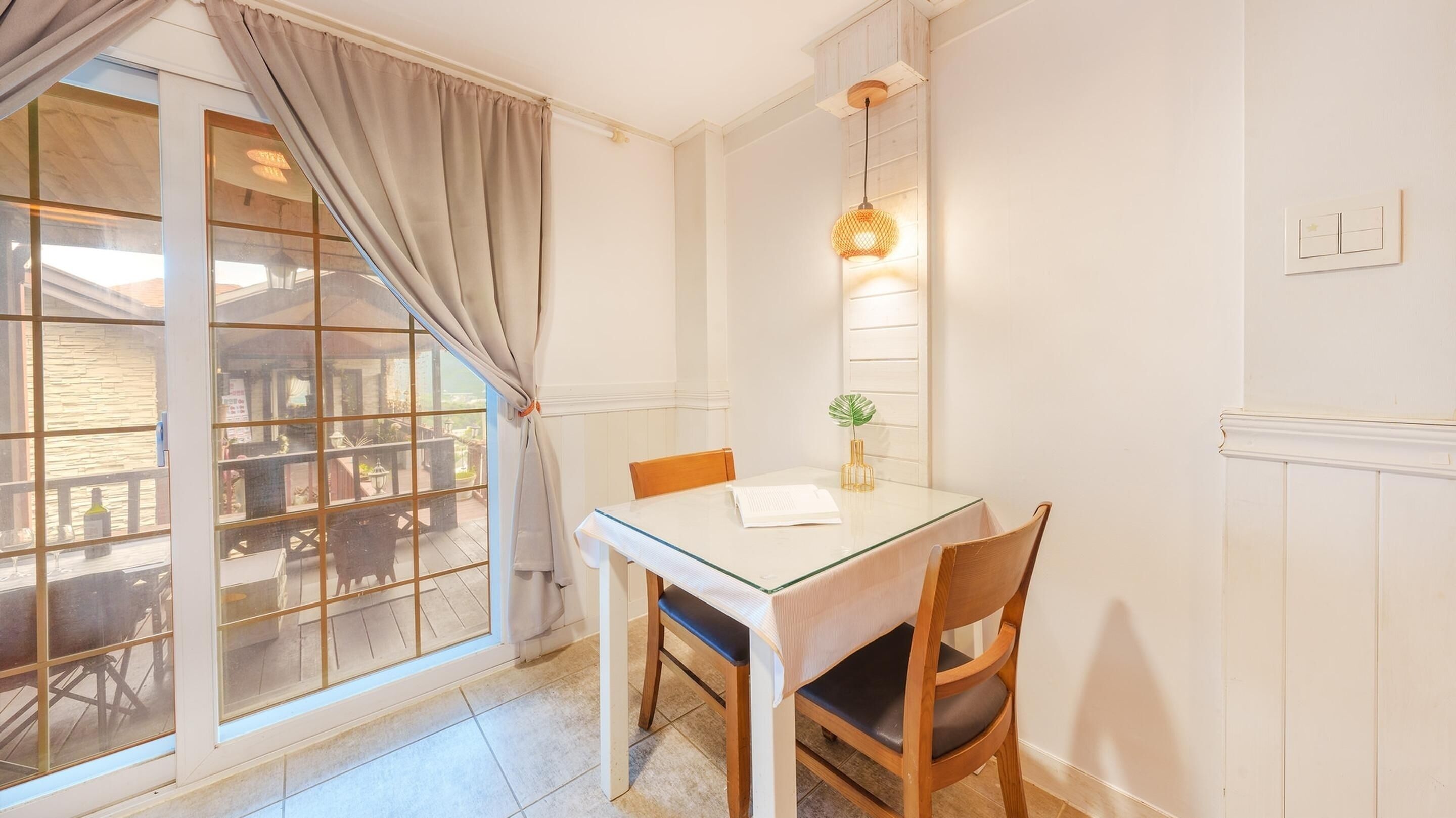 A dining area with a glass table, two chairs, a hanging light fixture, and a view of the outside through a window with curtains.
