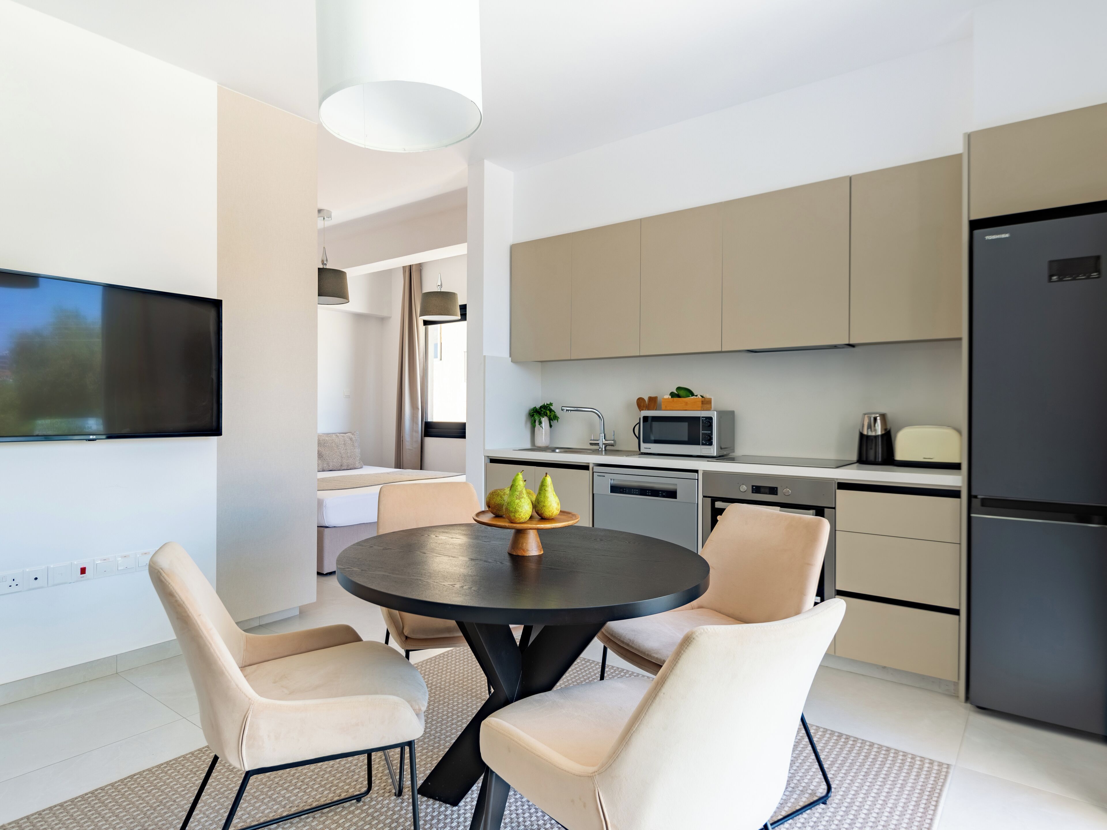 A modern kitchen with a dining area, including a round table with chairs, a flat-screen TV mounted on the wall, and a refrigerator.