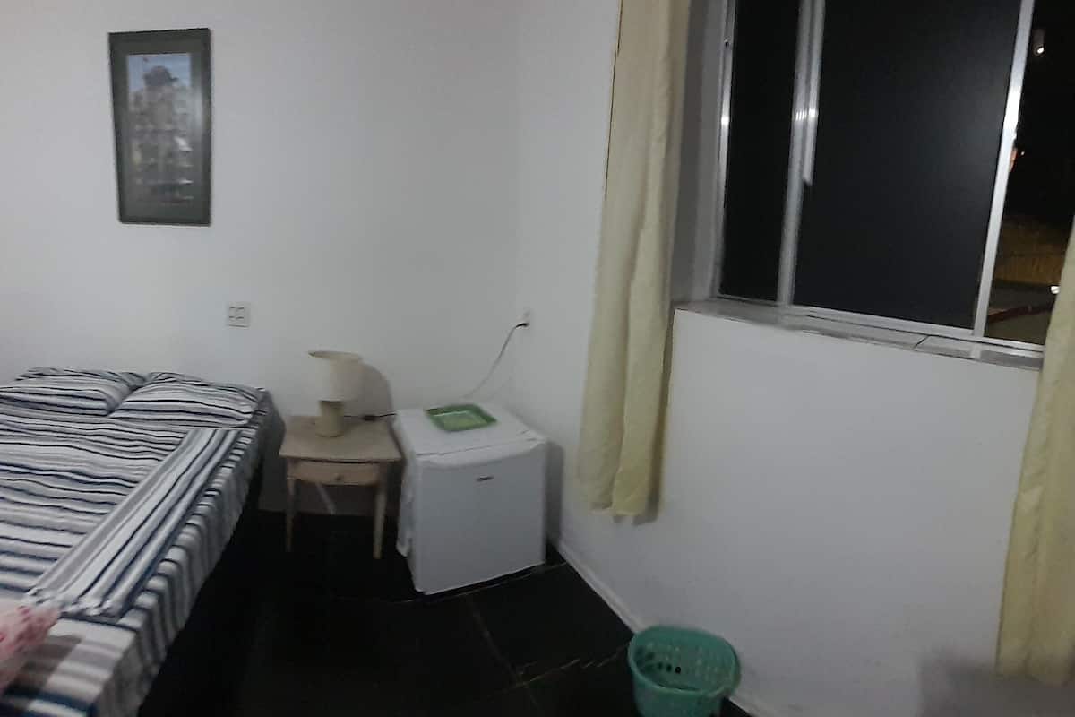 A small, sparsely furnished room with a single bed, a small table, a nightstand, a green trash bin, and a window with curtains.
