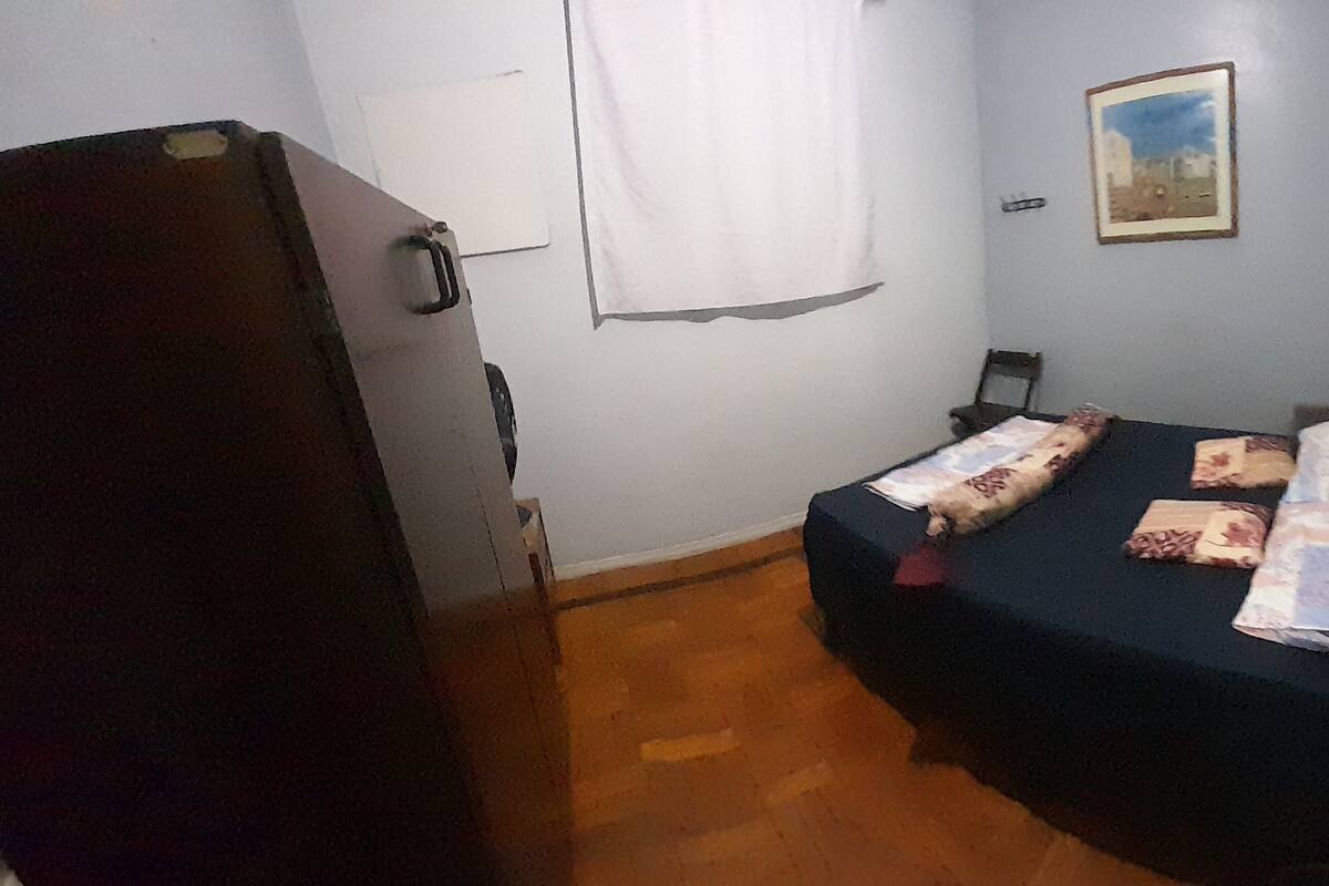 A small, sparsely furnished room with a single bed, a chair, a desk, and a wall-mounted picture.