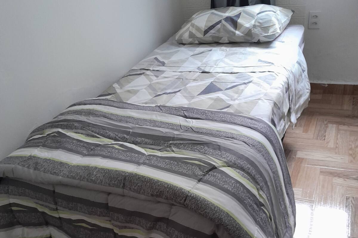 A small, neatly made bed with a patterned bedspread and pillows, a wooden floor, and a plain wall with a power outlet.