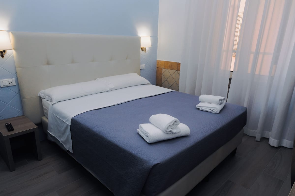 A neatly made bed with a blue bedspread and white linens, a bedside table with a remote control, and a wall-mounted lamp.
