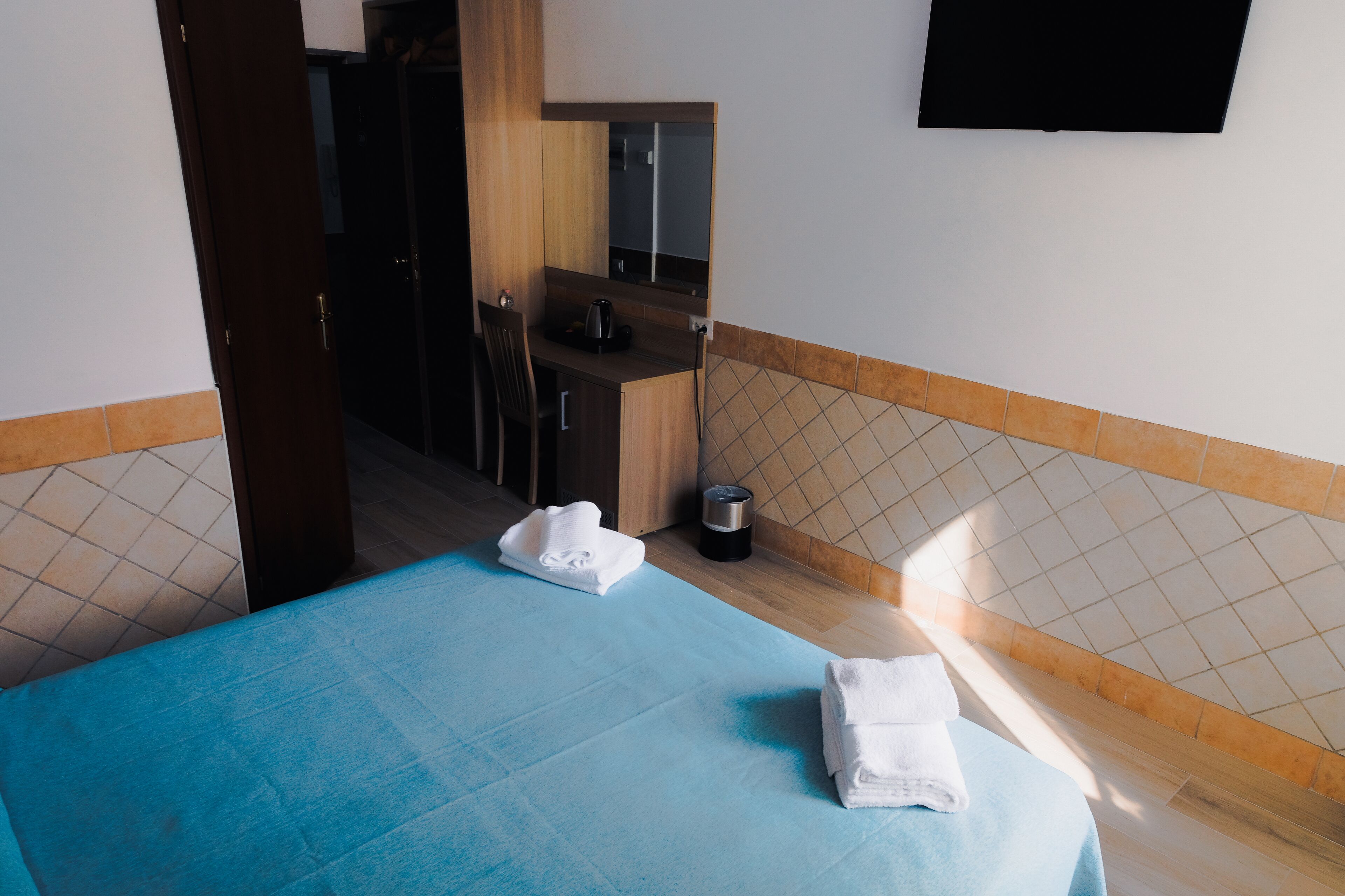Quarto de hotel com colcha azul, duas toalhas dobradas com cuidado, uma pequena mesa com chaleira e televisão fixada na parede.