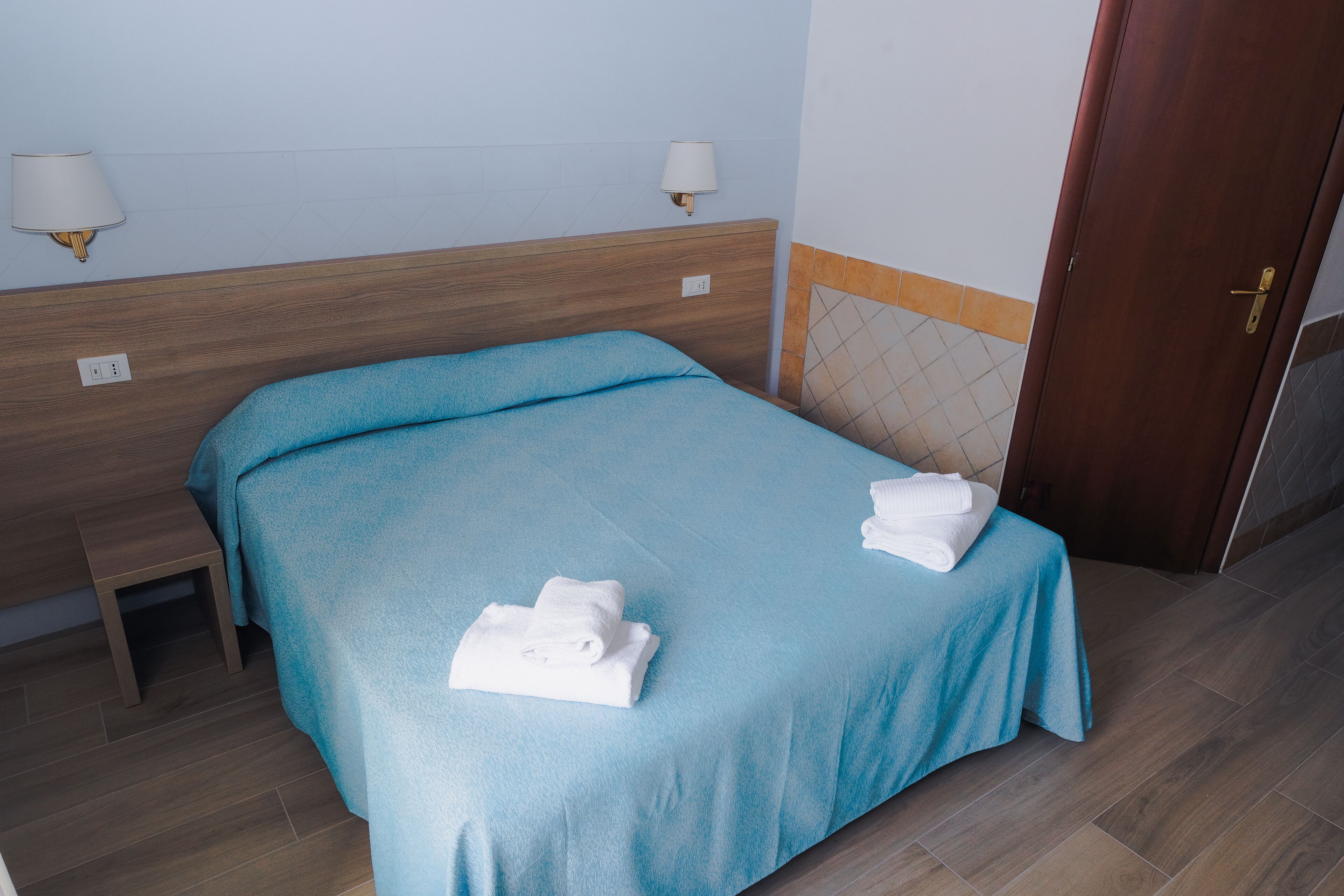 Una cama bien hecha con una colcha azul y toallas blancas, un cabecero de madera, una mesita de noche pequeña y una lámpara fijada a la pared.