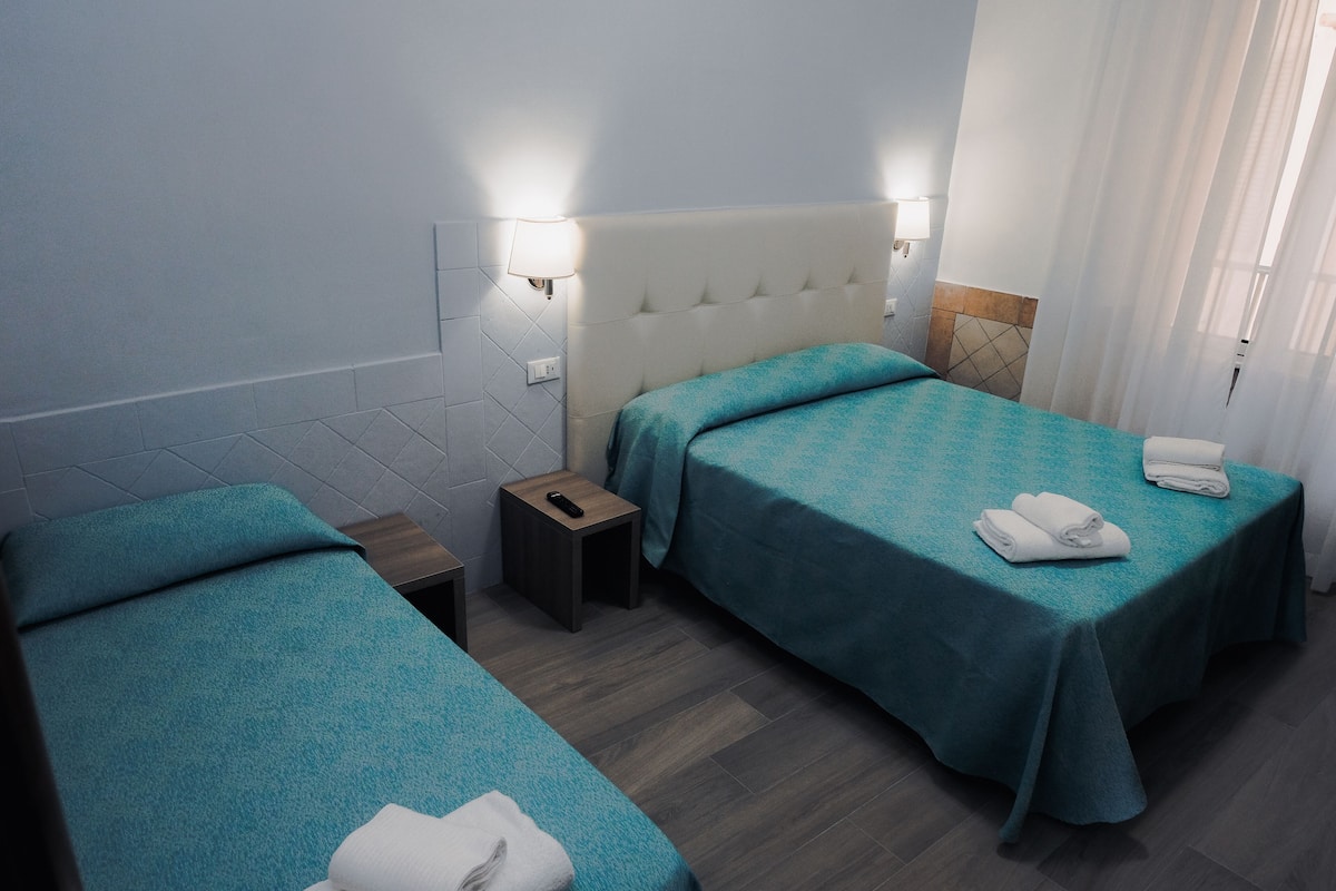 A hotel room with two single beds, a small bedside table, a nightstand, and a wall-mounted lamp.
