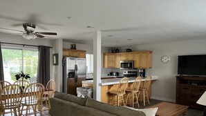 Een moderne keuken met roestvrijstalen apparatuur, een eethoek met houten stoelen, en een woonkamer met een bank en een televisie.