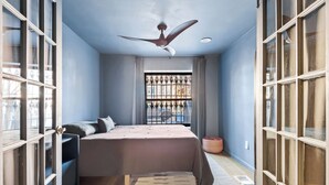 Une chambre à coucher avec un lit, un ventilateur de plafond, une fenêtre avec une grille en métal et une porte avec des panneaux de verre.
