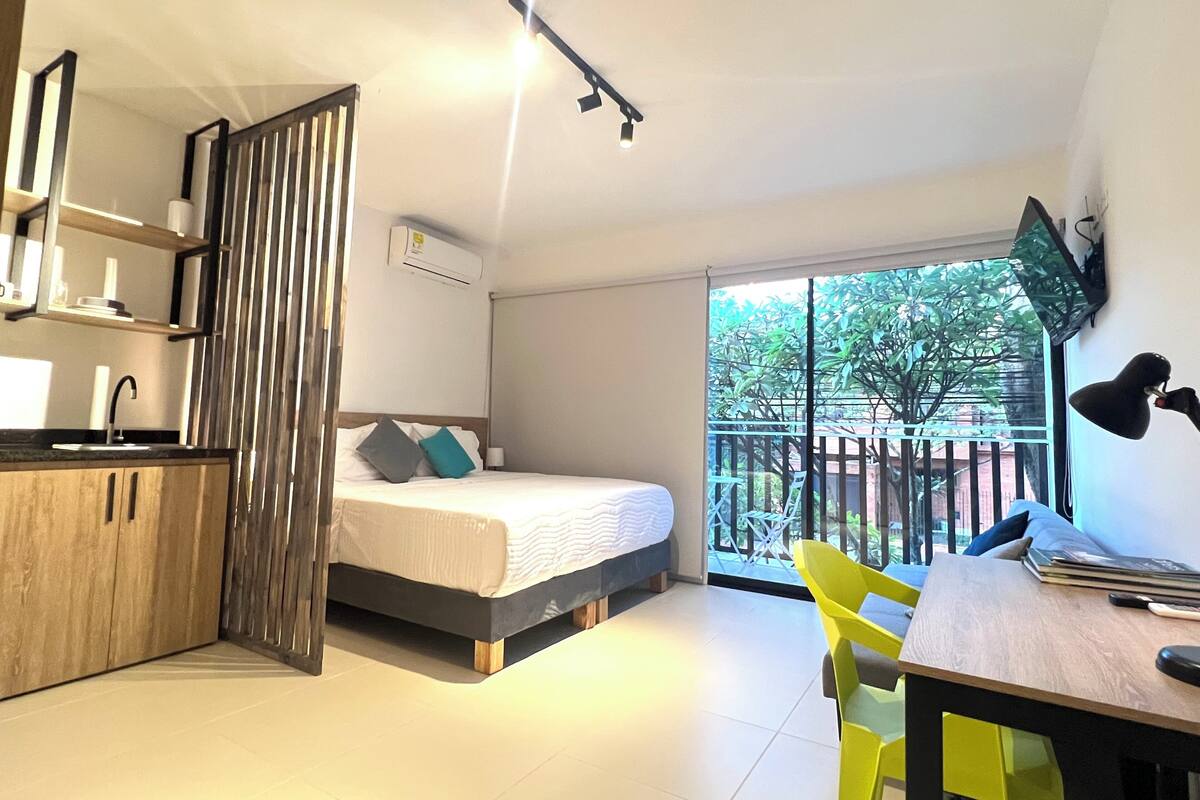 Una habitación moderna con cama, escritorio, silla, ventana con vista a follaje y una zona de cocina.