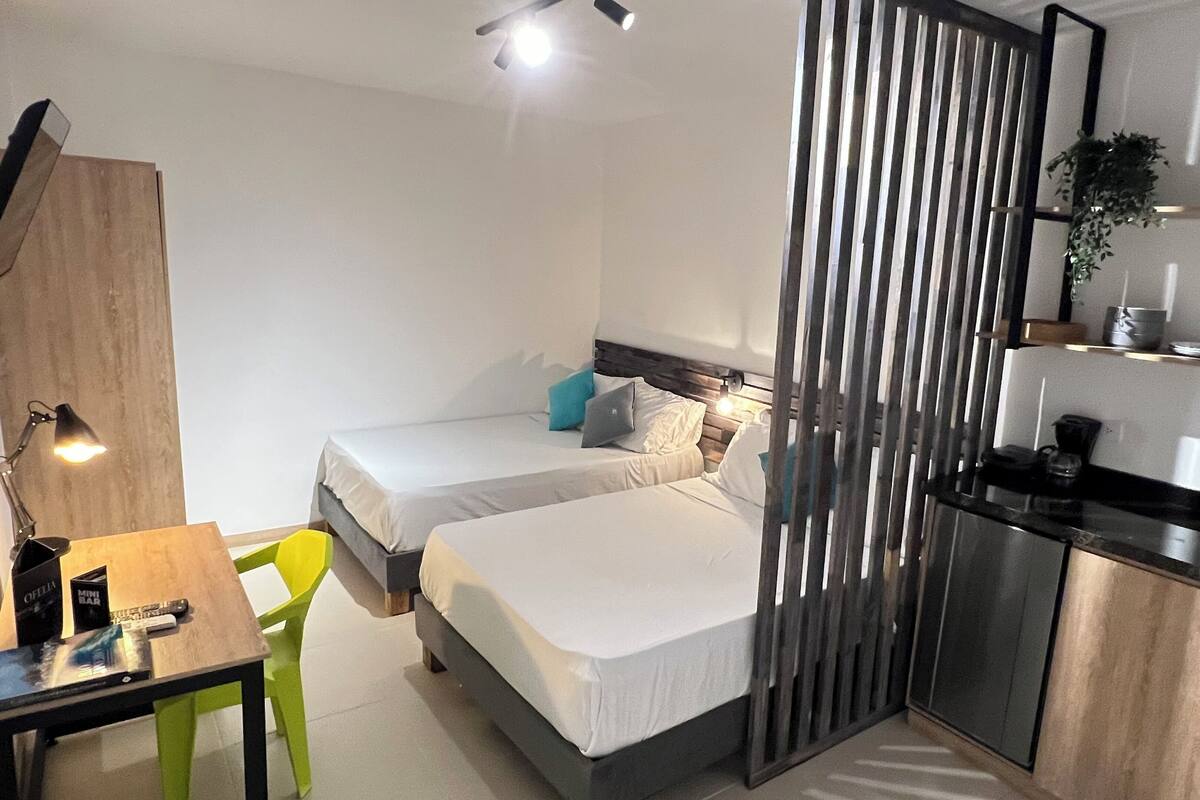 Una habitación de hotel compacta con dos camas individuales, un pequeño rincón americano, un escritorio con silla y un espejo.
