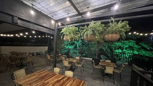 Een terras met houten tafels en stoelen, omringd door groen en verlicht met lichtslingers.