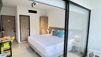 A modern hotel room with a large bed, a desk with a chair, a small table, and a trash bin.