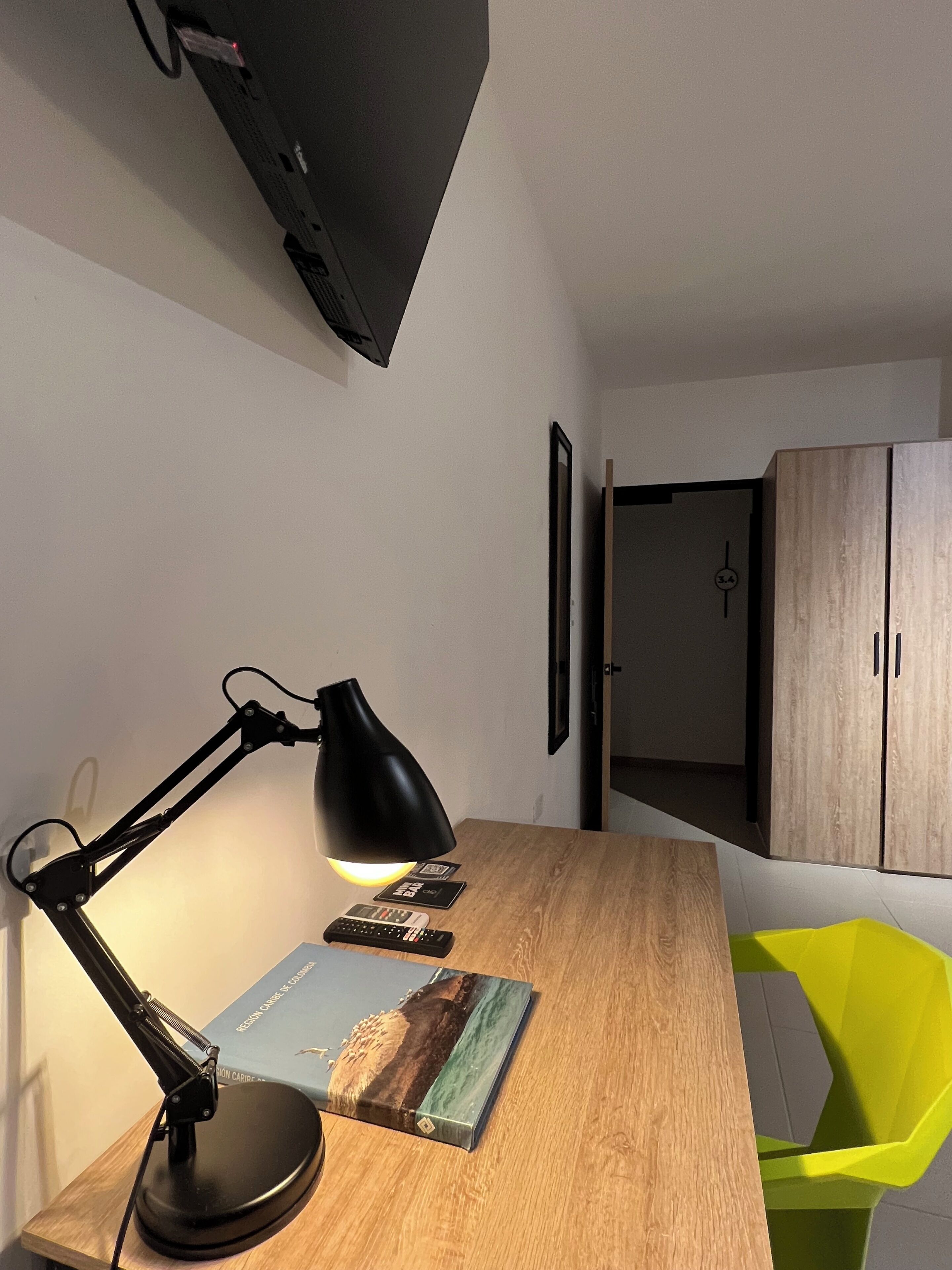 A desk with a desk lamp, a book, and remote controls. There is a wooden cabinet and a green chair in the background.