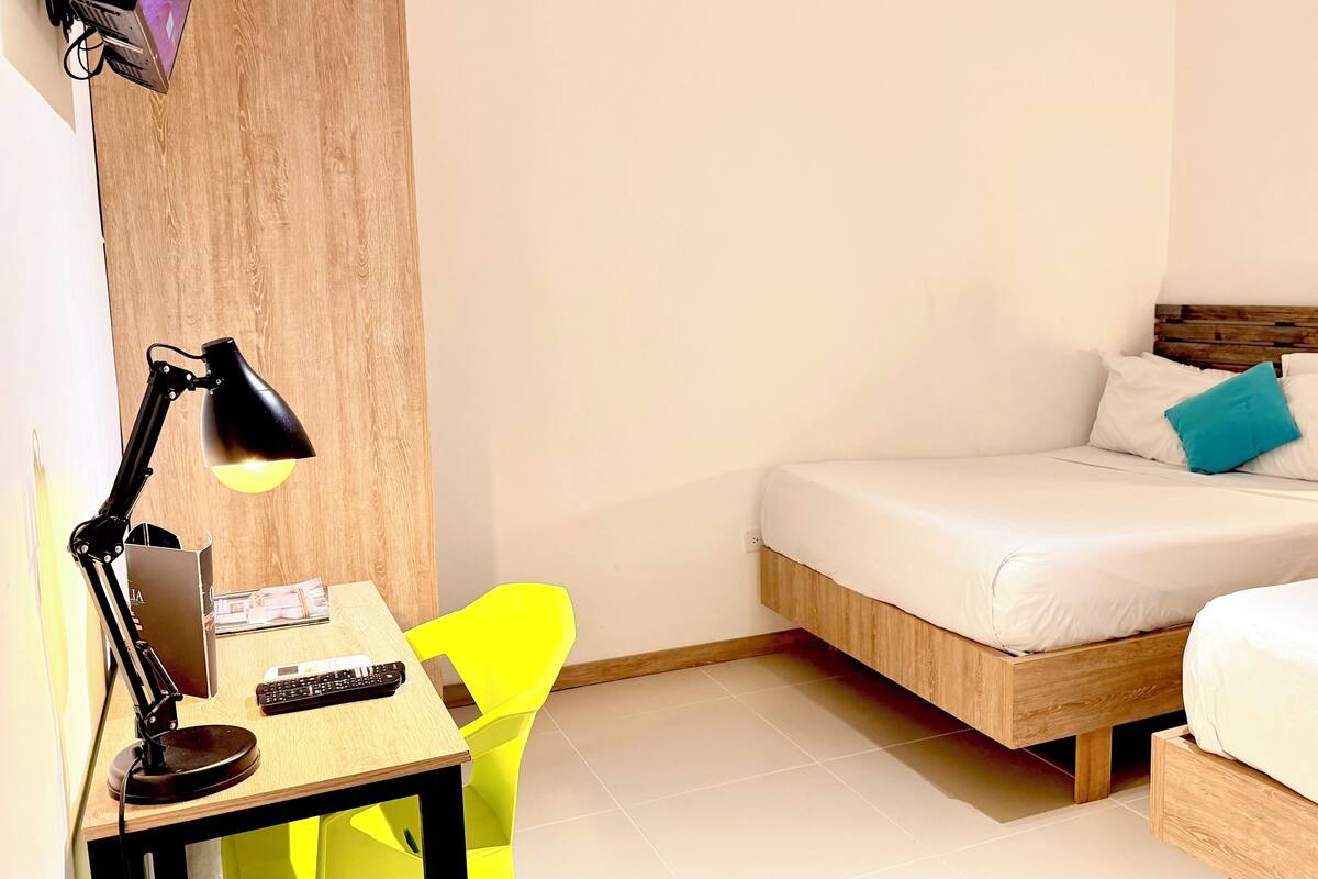 Una habitación de hotel pequeña con una cama de madera, un escritorio con lámpara, una silla y una mesita de noche.