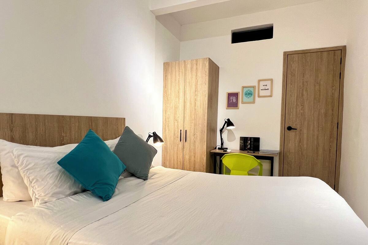 Una cama bien hecha con almohadas blancas y turquesa, un armario de madera, un escritorio con lámpara y una puerta.