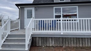 Une maison à deux étages avec une terrasse à l’avant, une porte blanche et une fenêtre donnant sur le ciel.