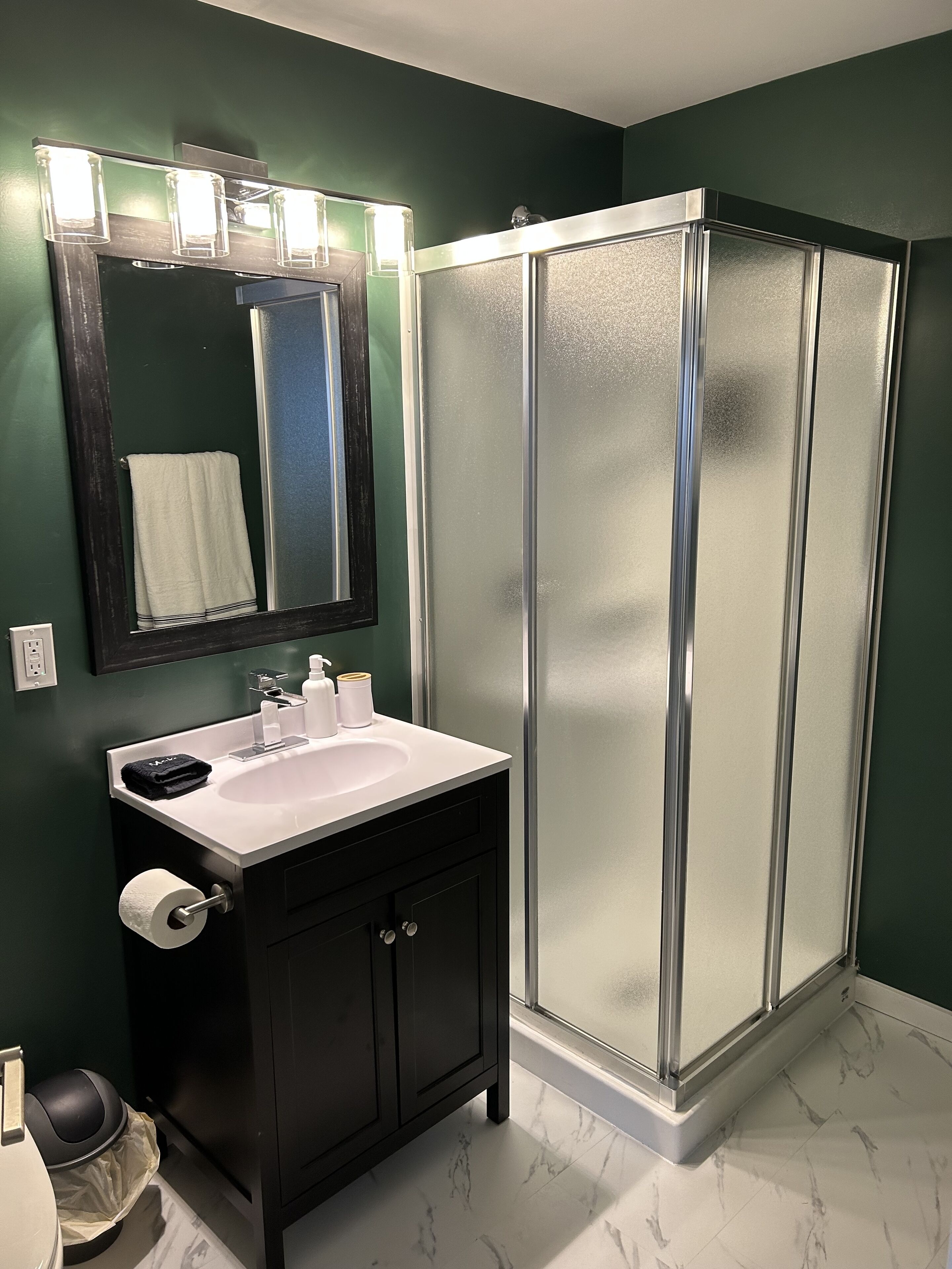 Un baño con un lavabo blanco, armarios oscuros, cabina de ducha de vidrio y un espejo con iluminación.