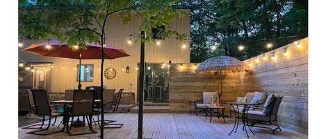 An outdoor patio with wooden flooring, a dining table with chairs, a red umbrella, and string lights.