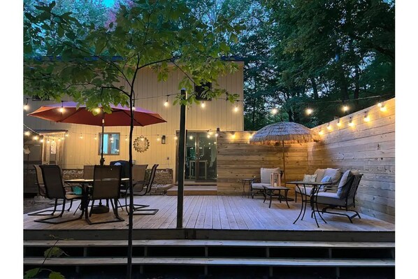 An outdoor patio with wooden flooring, a dining table with chairs, a red umbrella, and string lights. - Coastal theme/wooded & large deck/fenced-in yard (Holland)