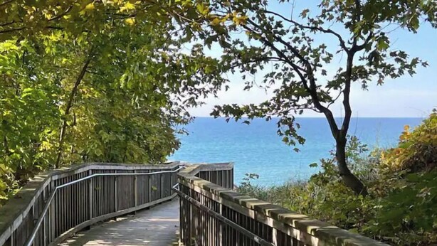 A wooden boardwalk with metal railings leading to a body of water. - Coastal theme/wooded & large deck/fenced-in yard (Holland)