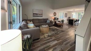 A spacious living room with a large sofa, a coffee table, and a ceiling fan. There is a dining area with a table and chairs visible through an open door. - Coastal theme/wooded & large deck/fenced-in yard (Holland)