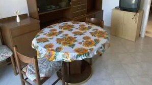 Una habitación con una mesa redonda cubierta por un mantel floreado, un armario, un televisor sobre un mueble bajo y una lámpara colgante.