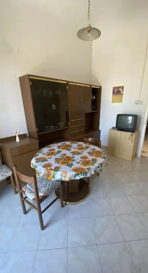 Una habitación con una mesa redonda cubierta por un mantel floreado, un armario, un televisor sobre un mueble bajo y una lámpara colgante.