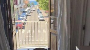 Un balcón con una mesa cubierta con mantel floreado, con vistas a una calle con coches aparcados y edificios.
