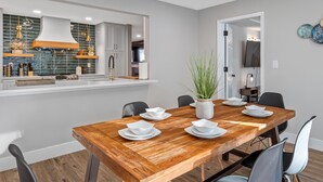 A modern dining area with a wooden table set for four, surrounded by black chairs. A potted plant is on the table, and the room has a neutral color scheme with a kitchen visible in the background.