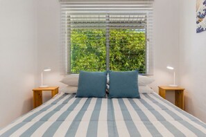 Een bed met gestreepte lakens, twee kussens, een raam met zonwering en twee nachtkastjes met lampen.