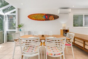 Uma sala de jantar com uma mesa de madeira, cadeiras brancas e uma obra de arte colorida na parede.
