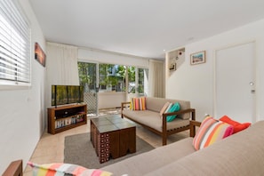 A living room with a sofa, a coffee table, a TV stand with books, and a sliding glass door leading to a balcony. - Kareela Court 4 Kareela Avenue 14 (Noosa Heads)