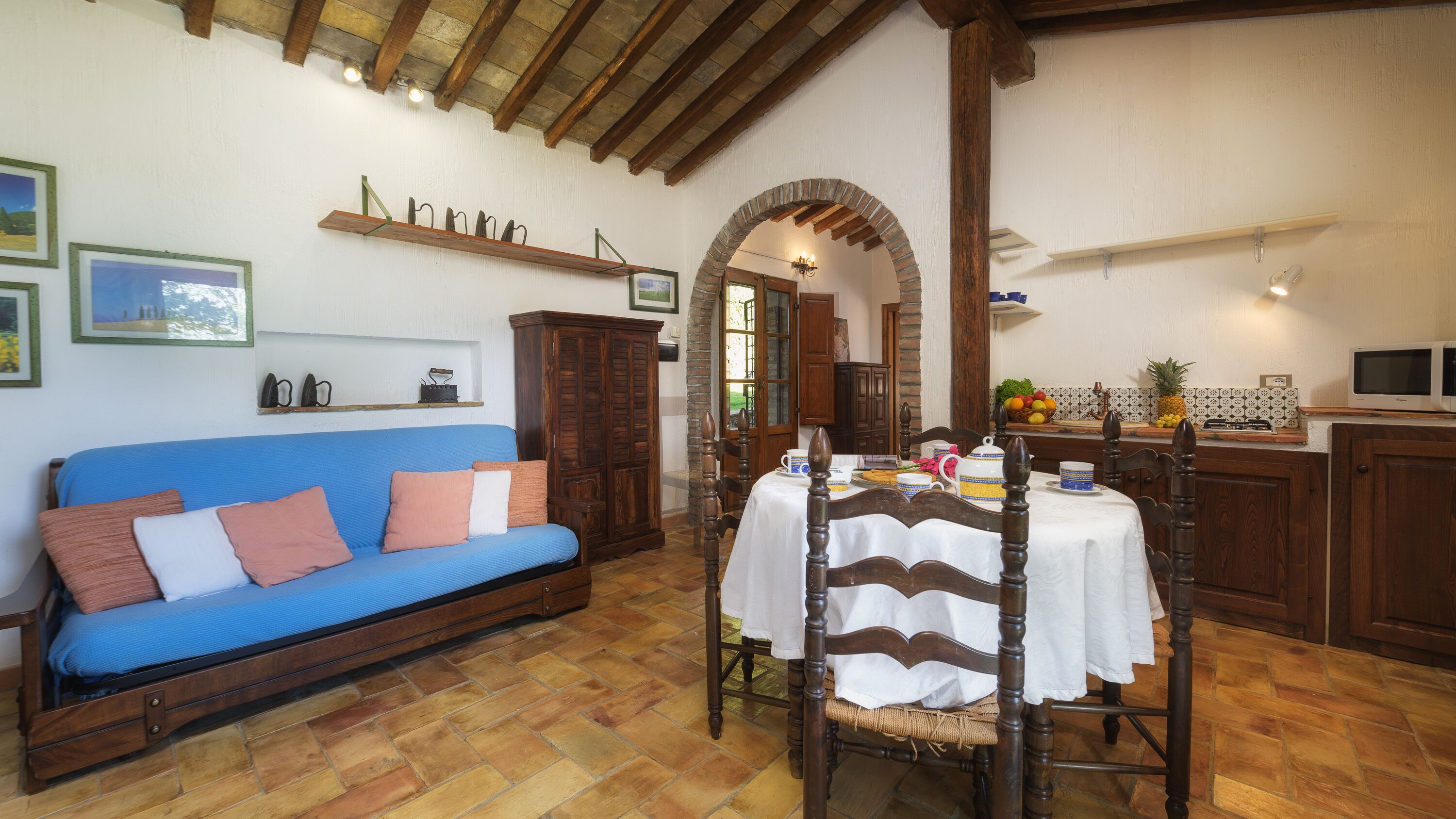 Una sala de estar acogedora con un sofá azul, una mesa de comedor con mantel blanco, sillas de madera y una zona de cocina con microondas.
