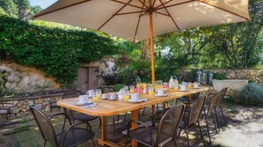 A wooden table set for breakfast with chairs, an umbrella, and a stone wall with greenery in the background.