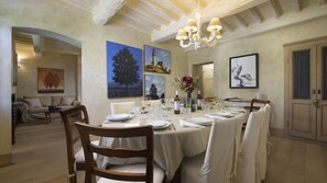 Comedor con una larga mesa dispuesta para seis personas, rodeada de sillas de madera. Hay pinturas en las paredes, un candelabro sobre la mesa y una puerta con pomo decorativo.