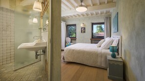 A bathroom with a glass shower enclosure, a sink, and a window. The bedroom features a bed, a nightstand, and a window with a view of trees.