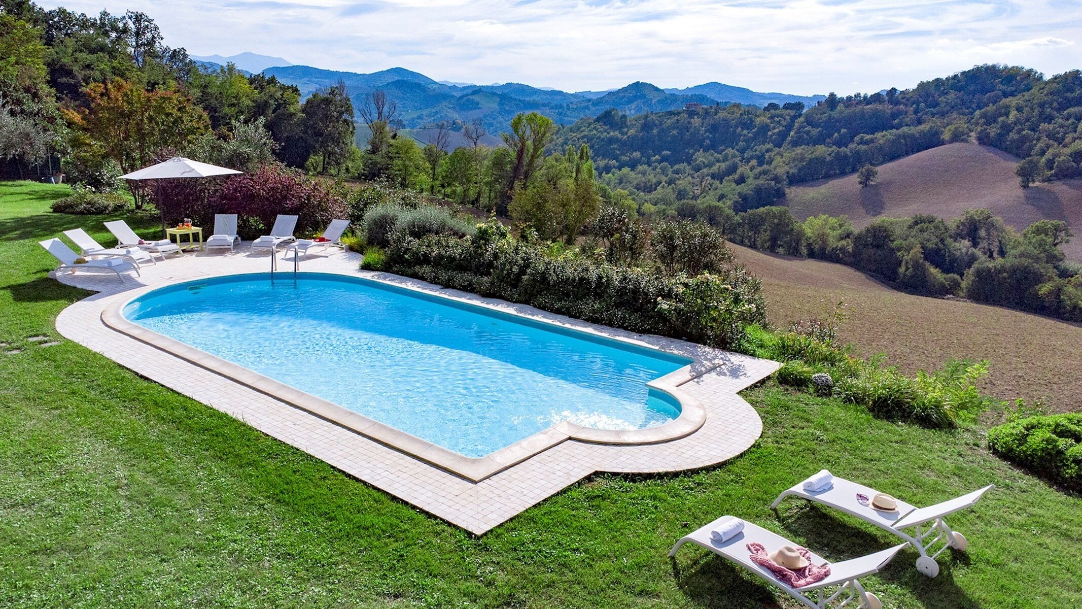 Une piscine rectangulaire, bordée d’une terrasse carrelée, entourée de pelouse et agrémentée de quelques chaises longues.