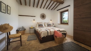 Een slaapkamer met stenen vloer, een bed met beddengoed met een patroon, een houten plafond en een raam met uitzicht.