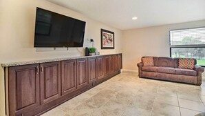 A modern kitchen with dark wooden cabinets, a large flat-screen TV mounted above the counter, and a beige tiled floor. - On-Site Pond: Sleek Largo Studio < 7 Mi to Beach! (Largo)