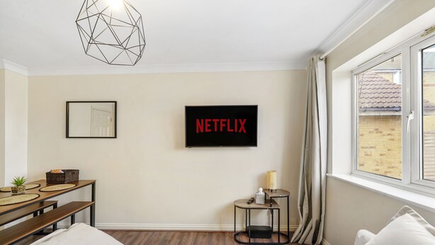 A bedroom with a bed, a desk with a chair, a small table with a lamp, a wall-mounted TV, and a window with curtains. - 4 bed Townhouse- Free parking (Greater London)