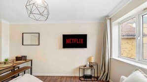 A hotel room with a bed, a desk with a lamp, a small table with a lamp, a wall-mounted TV, and a window with curtains.