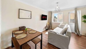 A living room with a sofa, a dining table with placemats, a wall-mounted TV, and a window with curtains.