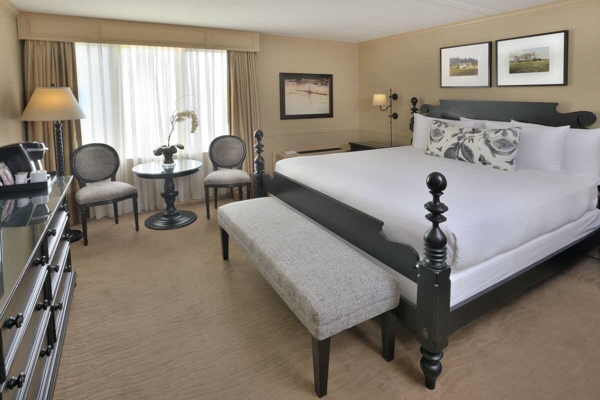 Ein Hotelzimmer mit einem großen Bett, einem Schreibtisch mit Lampe, zwei Stühlen, einem kleinen runden Tisch mit einer Vase und einer Bank.