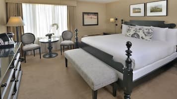 Une chambre d’hôtel avec un grand lit, un bureau avec une lampe, deux chaises, une petite table ronde avec un vase et un banc.