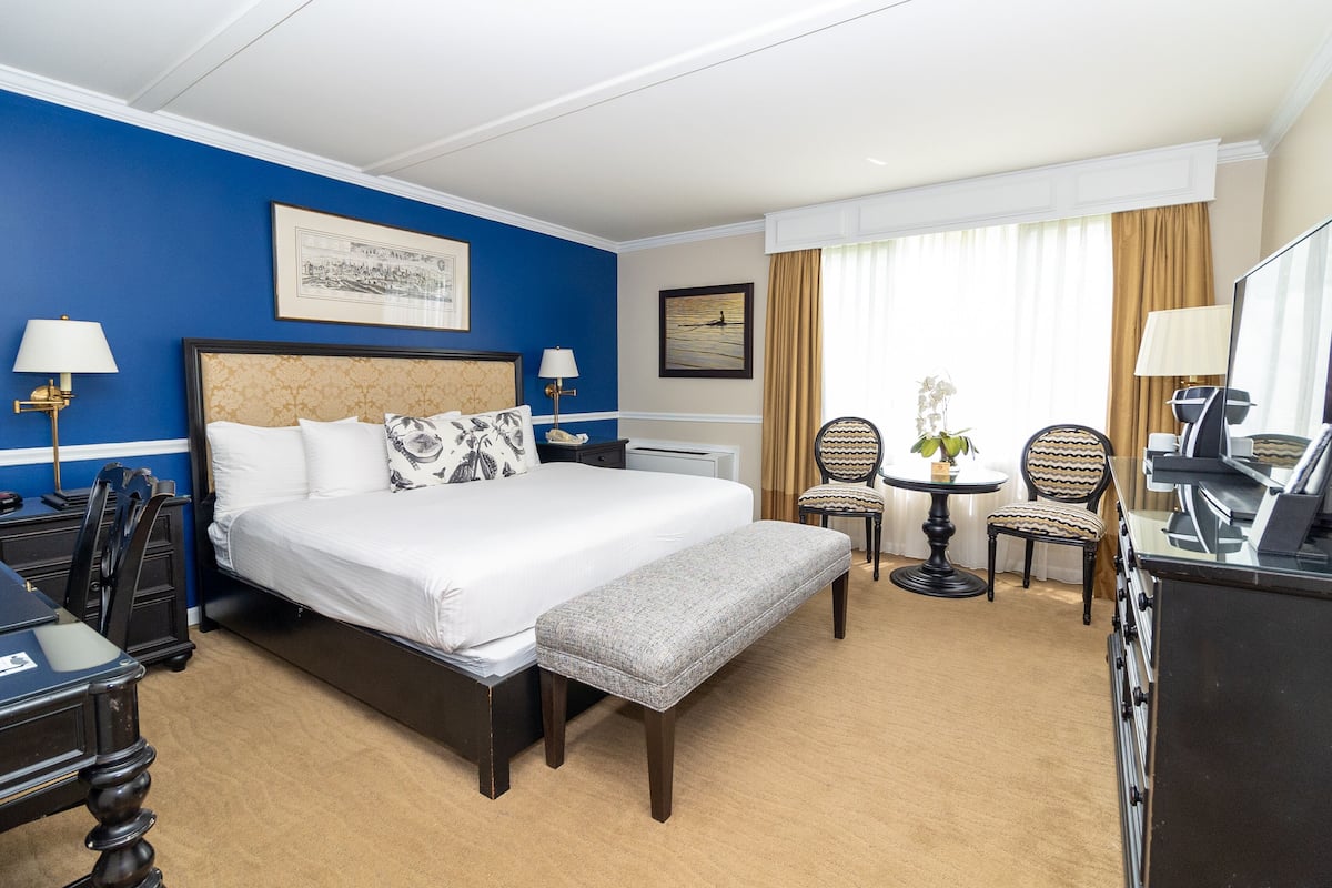 Ein Hotelzimmer mit einem großen Bett, einem Schreibtisch, zwei Stühlen, einem kleinen Tisch mit Lampe und einem Fernseher.