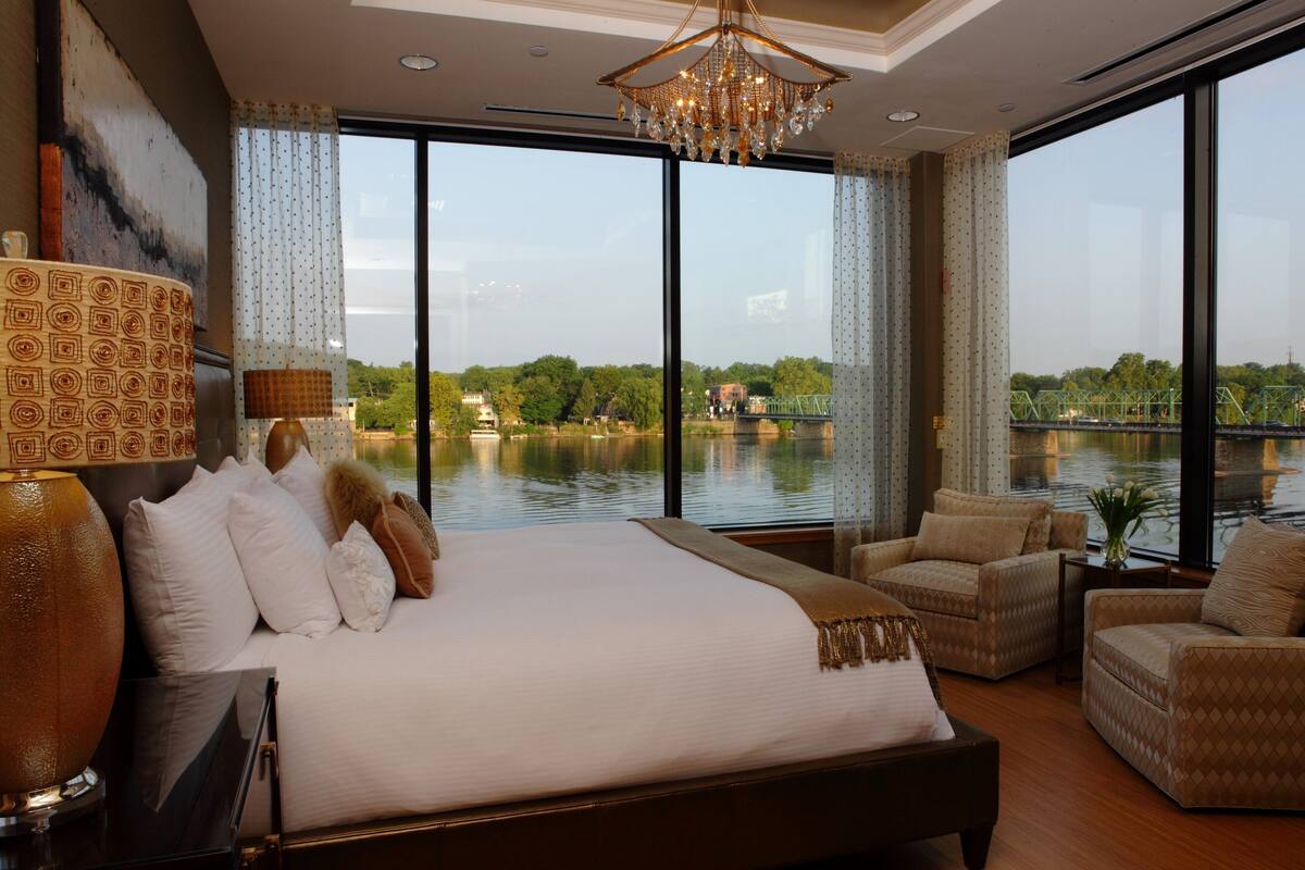 Ein geräumiges Schlafzimmer mit einem großen Bett, zwei Sesseln, einem Nachttisch und Blick auf einen Fluss und eine Brücke.