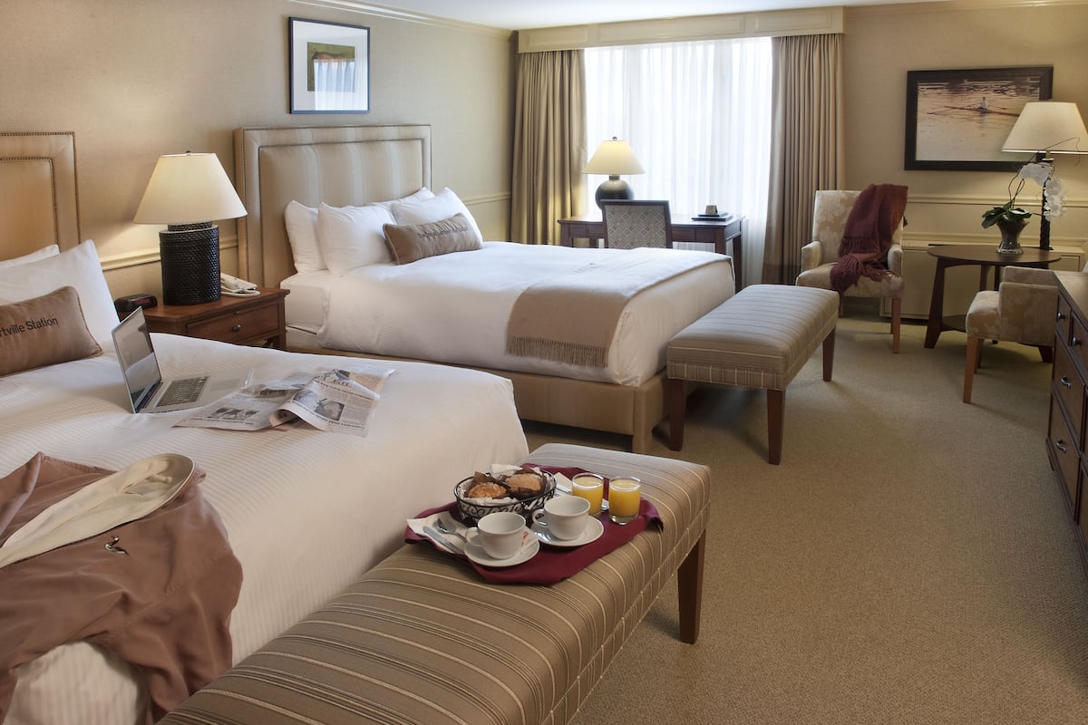 Ein Hotelzimmer mit zwei Betten, einem Schreibtisch, einem Stuhl und einem kleinen Tisch mit einer Lampe.
